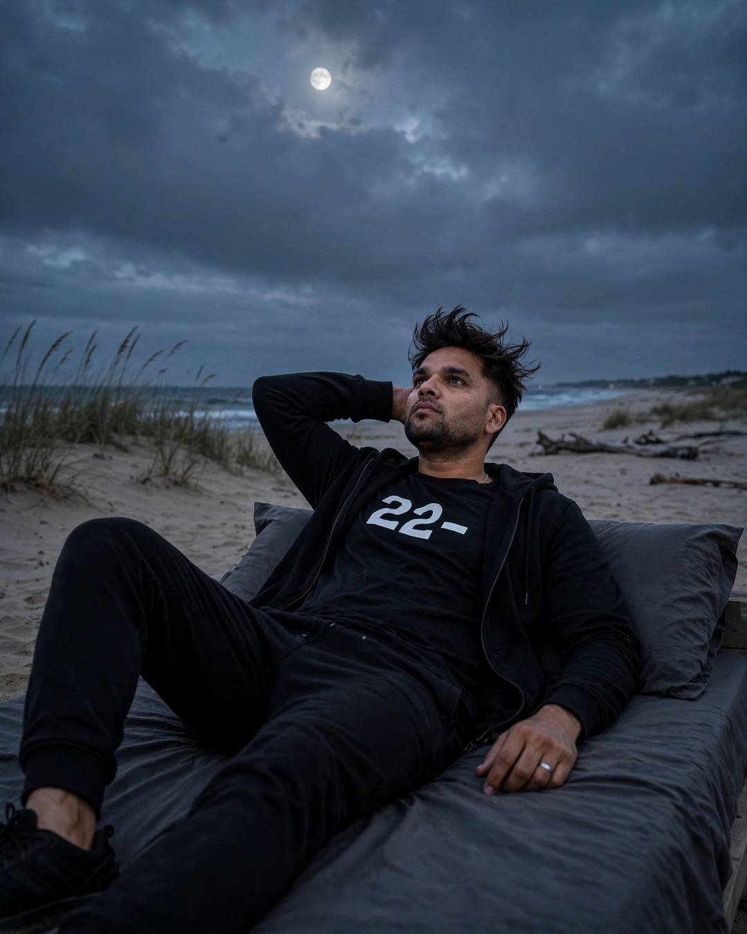 A young Indian man in all-black streetwear reclining on a bed on a deserted beach at dusk, gazing at the moon through heavy clouds